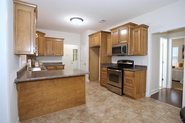 kitchen featuring a sink, dark countertops, appliances with stainless steel finishes, a peninsula, and brown cabinetry