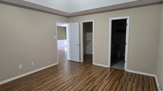 unfurnished bedroom with dark wood-style floors, baseboards, a closet, ensuite bathroom, and a walk in closet