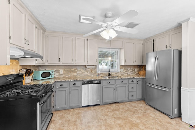 kitchen with gray cabinetry, under cabinet range hood, a sink, appliances with stainless steel finishes, and light stone countertops