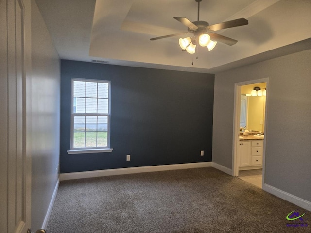 unfurnished bedroom featuring ceiling fan, baseboards, a tray ceiling, carpet floors, and ensuite bath