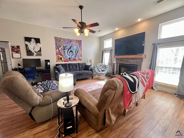 living area with visible vents, a textured ceiling, wood finished floors, a stone fireplace, and ceiling fan