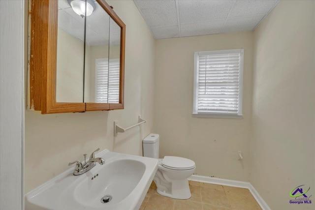 half bath with tile patterned flooring, toilet, baseboards, a drop ceiling, and a sink