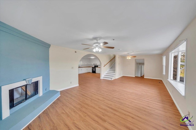 unfurnished living room with stairs, a fireplace with raised hearth, arched walkways, light wood-style floors, and a ceiling fan