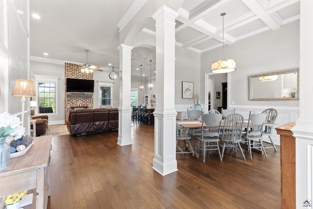 dining area featuring dark wood-style flooring, a brick fireplace, beam ceiling, ceiling fan, and decorative columns