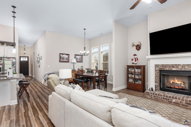 living room with baseboards, wood finished floors, ceiling fan with notable chandelier, and a brick fireplace