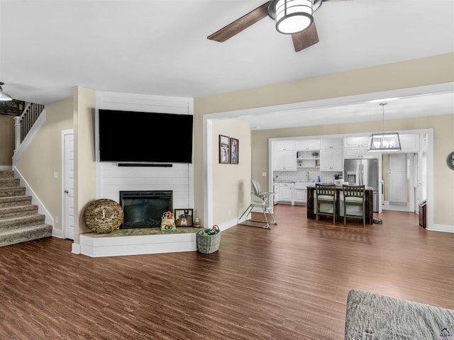 living area with ceiling fan, a fireplace, dark wood finished floors, stairs, and baseboards