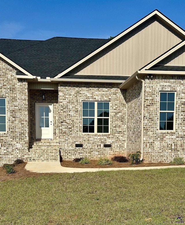 view of exterior entry featuring brick siding, a shingled roof, a lawn, and crawl space