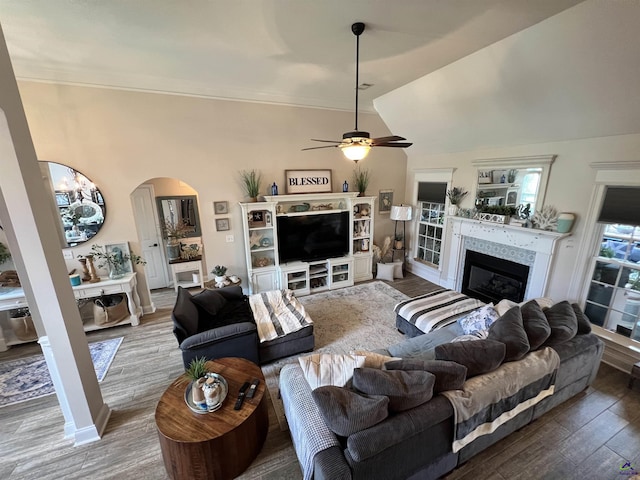 living area with arched walkways, ceiling fan, wood finished floors, a glass covered fireplace, and crown molding