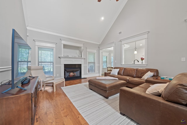 living area with high vaulted ceiling, crown molding, light wood-style flooring, a lit fireplace, and a ceiling fan