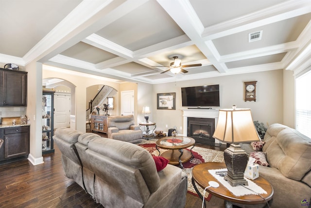 living room featuring arched walkways, coffered ceiling, dark wood finished floors, beam ceiling, and a warm lit fireplace