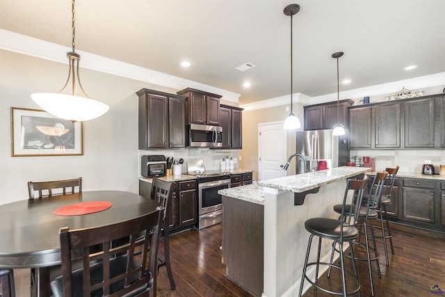 kitchen featuring appliances with stainless steel finishes, ornamental molding, dark brown cabinets, tasteful backsplash, and an island with sink