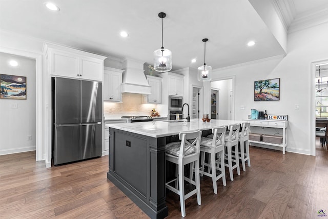 kitchen with stainless steel appliances, custom exhaust hood, white cabinets, crown molding, and a center island with sink