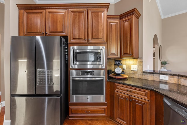 kitchen featuring appliances with stainless steel finishes, crown molding, backsplash, dark stone countertops, and brown cabinets