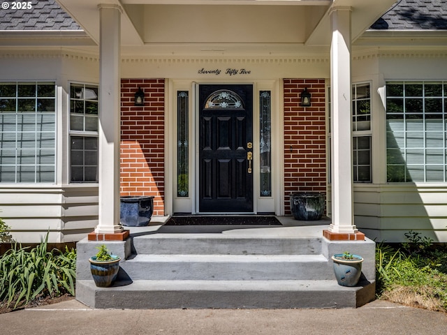 view of exterior entry with covered porch, brick siding, and roof with shingles
