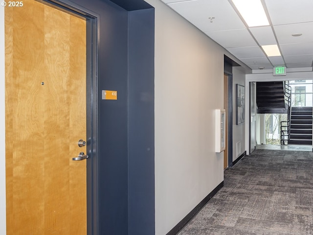 corridor featuring stairway, dark carpet, a paneled ceiling, and baseboards