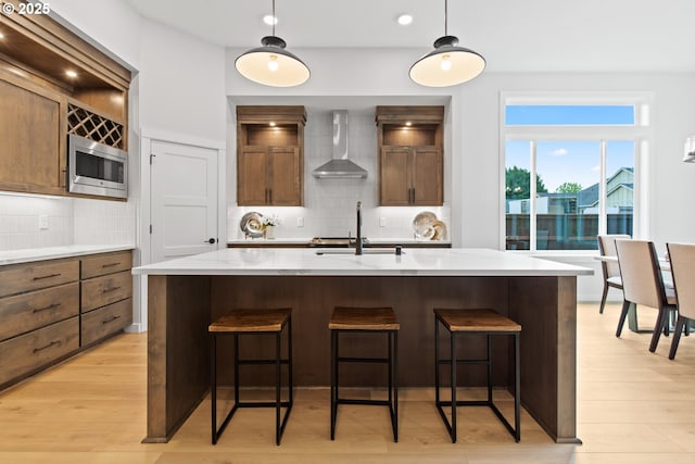 kitchen featuring light wood-style flooring, a kitchen bar, stainless steel microwave, wall chimney exhaust hood, and a sink