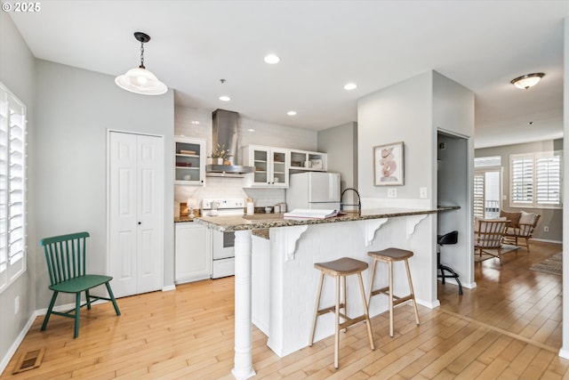 kitchen with a breakfast bar, light wood-style floors, wall chimney range hood, white appliances, and a peninsula