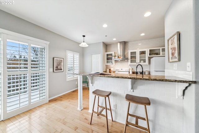 kitchen featuring light wood finished floors, a kitchen breakfast bar, freestanding refrigerator, a peninsula, and wall chimney range hood