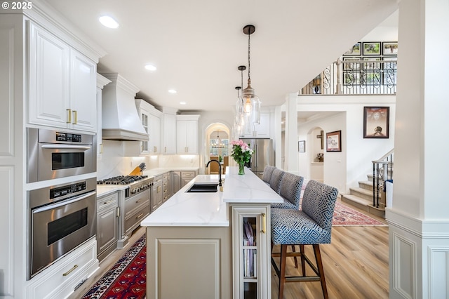 kitchen with arched walkways, custom range hood, stainless steel appliances, a kitchen bar, and a sink