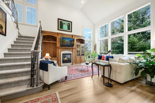 living room featuring high vaulted ceiling, a glass covered fireplace, wood-type flooring, and stairs