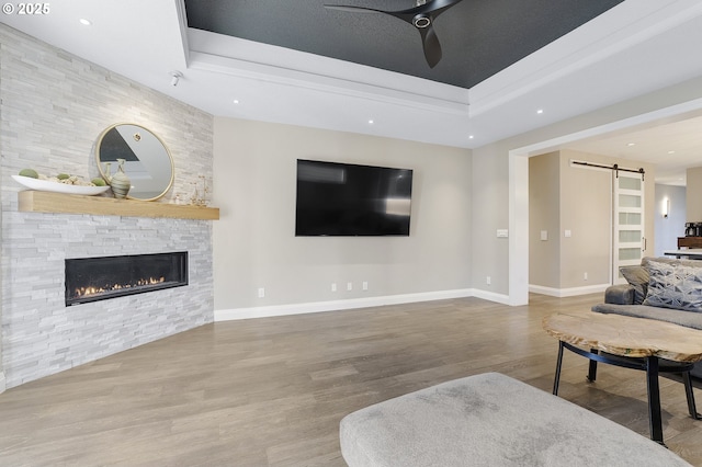 living room featuring ceiling fan, a barn door, a tray ceiling, hardwood / wood-style flooring, and a stone fireplace