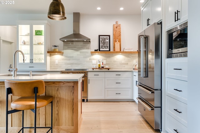 kitchen with high end appliances, wall chimney range hood, a sink, light wood-type flooring, and light countertops