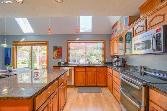 kitchen featuring brown cabinetry, light wood finished floors, a sink, appliances with stainless steel finishes, and vaulted ceiling with skylight