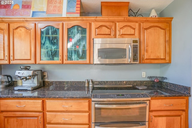 kitchen featuring brown cabinetry, dark stone countertops, appliances with stainless steel finishes, and glass insert cabinets