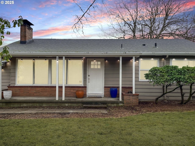 view of front of home with brick siding, roof with shingles, covered porch, a lawn, and a chimney