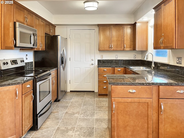 kitchen featuring a sink, dark stone counters, appliances with stainless steel finishes, and brown cabinetry