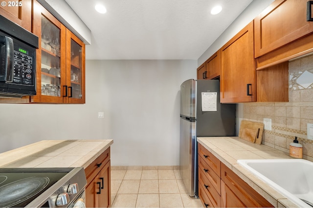 kitchen with brown cabinets, stainless steel appliances, glass insert cabinets, tasteful backsplash, and tile countertops