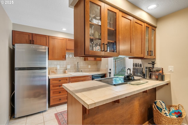 kitchen featuring freestanding refrigerator, tile counters, brown cabinets, backsplash, and a sink