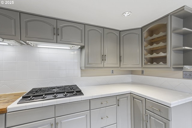 kitchen with gray cabinets, tile counters, ventilation hood, stainless steel gas cooktop, and decorative backsplash