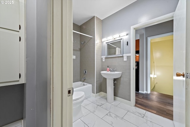 bathroom with tile walls, toilet, marble finish floor, washtub / shower combination, and a sink