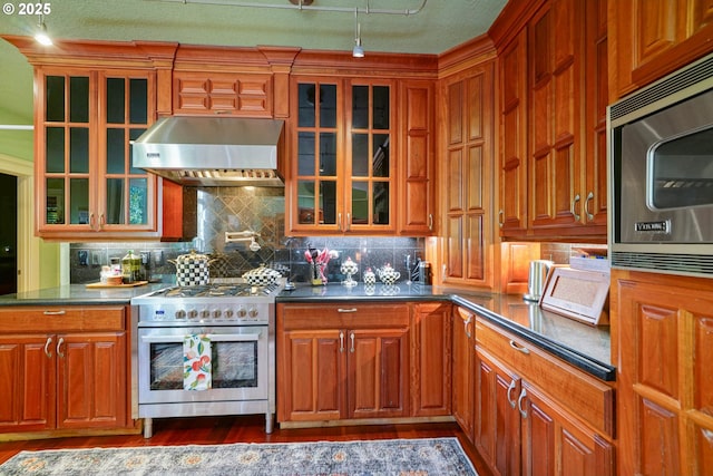 kitchen with tasteful backsplash, dark countertops, appliances with stainless steel finishes, glass insert cabinets, and exhaust hood