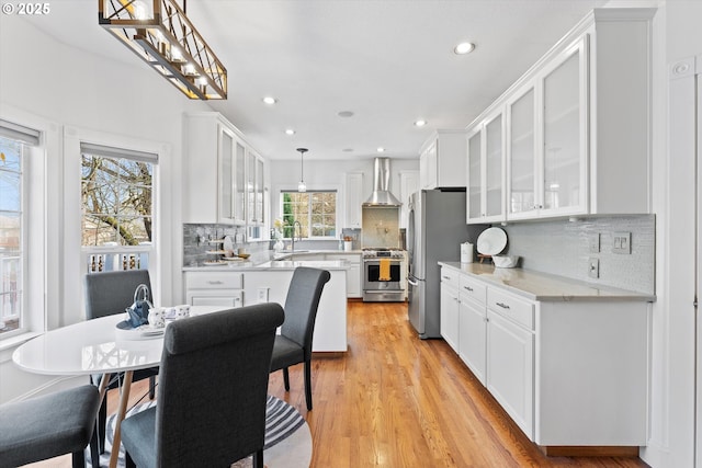 kitchen featuring pendant lighting, wall chimney range hood, white cabinetry, stainless steel appliances, and light stone counters