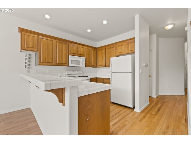 kitchen featuring brown cabinetry, a peninsula, white appliances, light wood-style floors, and tile counters