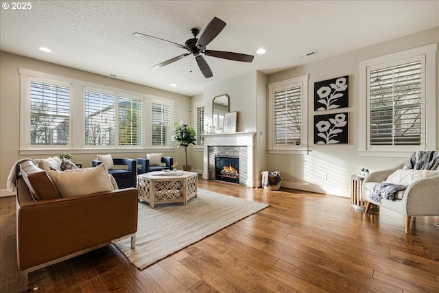 living room with wood-type flooring, a textured ceiling, a stone fireplace, and ceiling fan