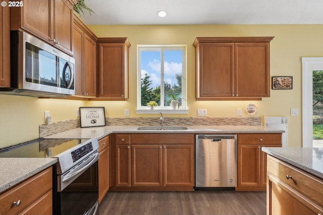 kitchen with brown cabinetry, dark wood-style floors, light stone counters, a sink, and stainless steel appliances