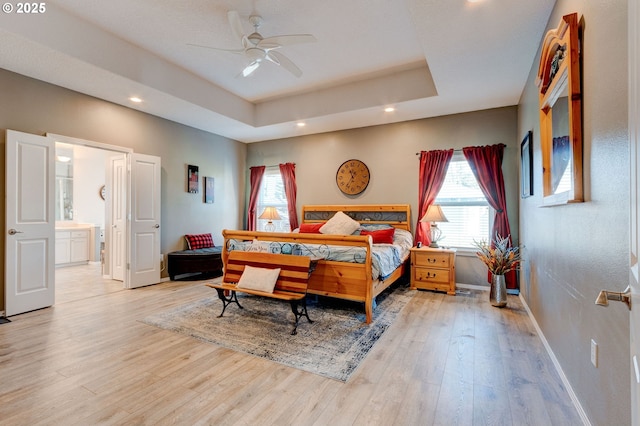 bedroom with ceiling fan, ensuite bath, a tray ceiling, and light hardwood / wood-style floors