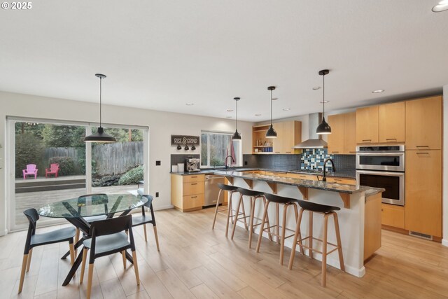 kitchen with pendant lighting, appliances with stainless steel finishes, light brown cabinetry, and wall chimney range hood