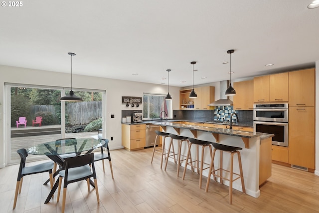 kitchen with a center island with sink, wall chimney exhaust hood, light brown cabinets, and stainless steel appliances