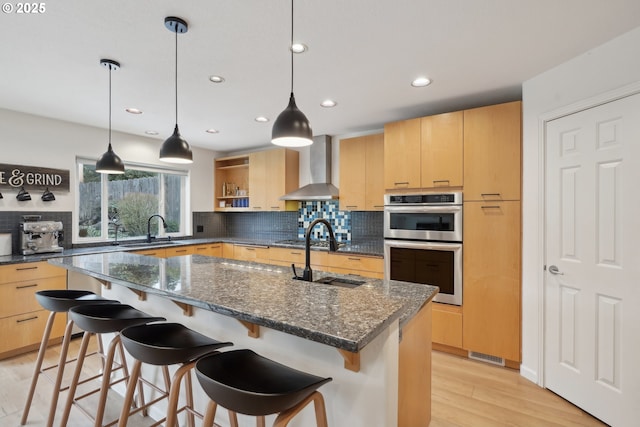 kitchen with a sink, double oven, wall chimney exhaust hood, and light brown cabinetry