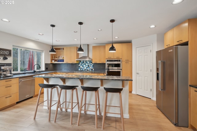 kitchen featuring light brown cabinets, wall chimney range hood, light wood-style floors, a kitchen bar, and stainless steel appliances