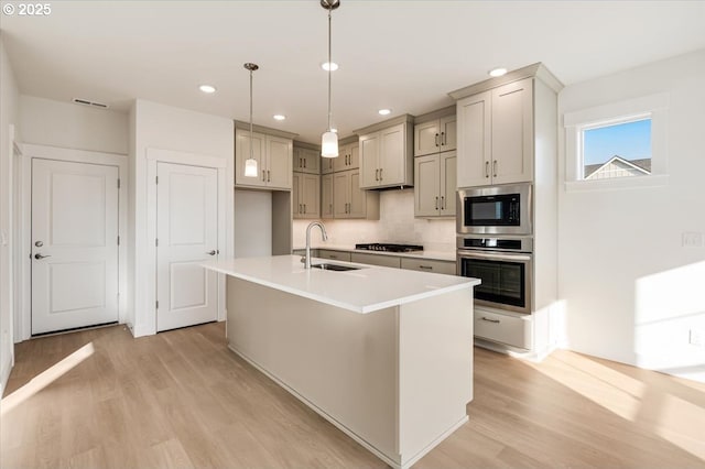 kitchen with sink, stainless steel appliances, light hardwood / wood-style floors, pendant lighting, and a kitchen island with sink