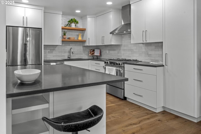 kitchen with stainless steel appliances, dark countertops, a sink, wall chimney range hood, and light wood-type flooring