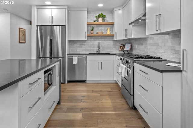 kitchen with light wood finished floors, dark countertops, appliances with stainless steel finishes, a sink, and wall chimney range hood