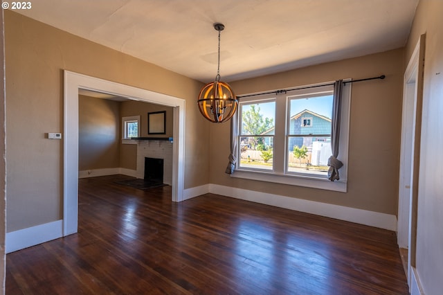 unfurnished dining area with a brick fireplace, dark wood-style floors, a notable chandelier, and baseboards