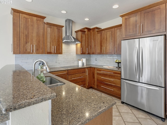 kitchen with freestanding refrigerator, wall chimney range hood, a sink, brown cabinets, and recessed lighting