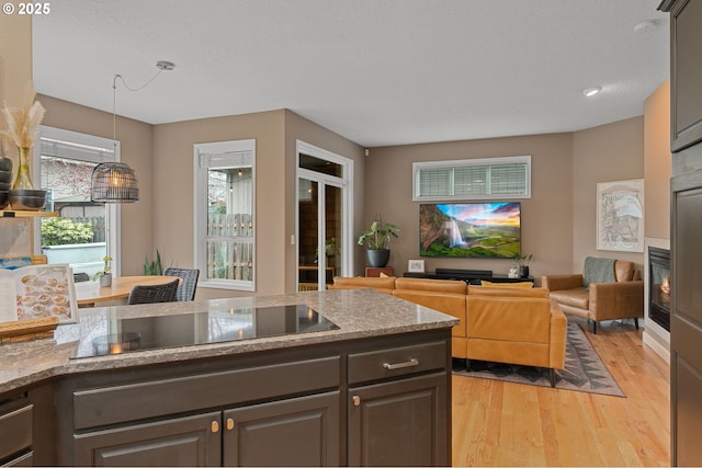 kitchen with pendant lighting, light wood-style flooring, a glass covered fireplace, black electric cooktop, and light stone countertops